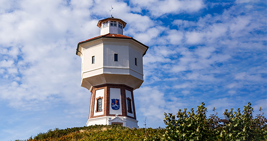 Das Foto zeigt einen eckigen Leuchtturm auf einer grünern Weise. Im Hintergrund ist der Himmel blau und bewölkt.