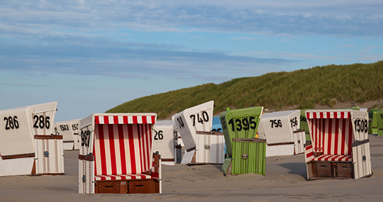 Das Foto zeigt einen Strand mit bunten Strandkörben. Im Hintergrund ist eine hohe, grün bewachsene Düne zu sehen. Der Himmel ist  blau.