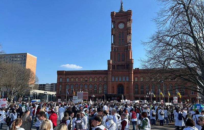 Das Bild zeigt eine große Menschenmenge auf einem Platz. Einige Personen halten Protestschilder hoch. Im Hintergrund ist ein rotes Gebäude mit einem roten Turm zu sehen.