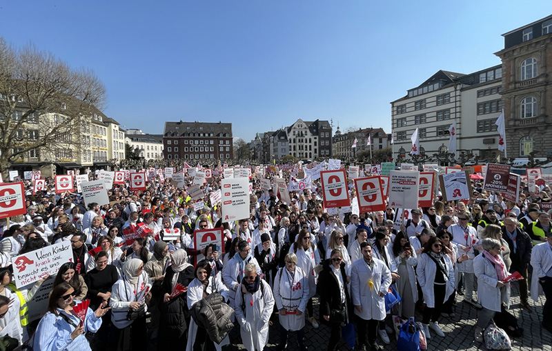 Das Bild zeigt eine große Menschenmenge auf einem Platz. Einige Personen halten Protestschilder hoch.