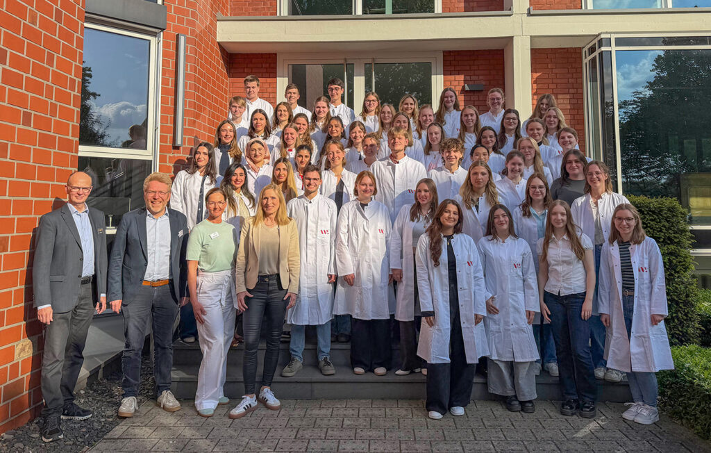 Gruppenfoto von Personen vor einem Backsteingebäude, viele tragen weiße Kittel, einige wenige tragen legere Kleidung.