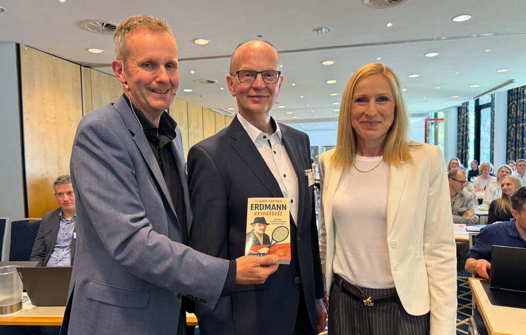 Gruppenbild Schmitz Erdmann Prinz Kv 1 12026 Drei Personen stehen nebeneinander in einem Raum mit Publikum im Hintergrund, eine Person hält ein Buch mit dem Titel 'ERDMANN ermittelt'