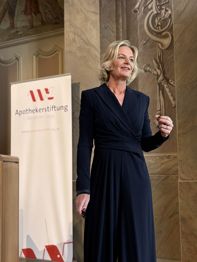 18_stiftungsveranstaltung_aswl_lioba_werth_web Frau in schwarzem Kleid steht vor einer Wand mit Relief und einem Banner der Apothekerstiftung Westfalen-Lippe