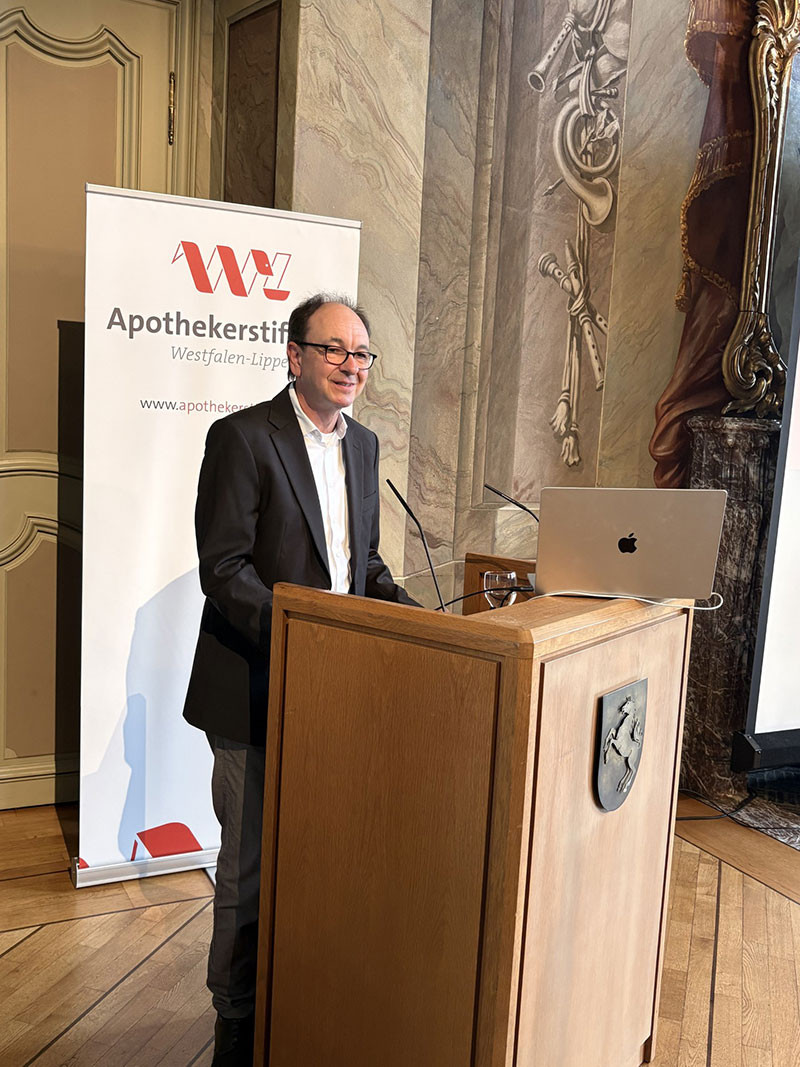 Ein Mann mit Brille und dunklem Sakko steht an einem Rednerpult vor einem Banner der Apothekerstiftung Westfalen-Lippe und einem geöffneten Laptop in einem prunkvollen Raum